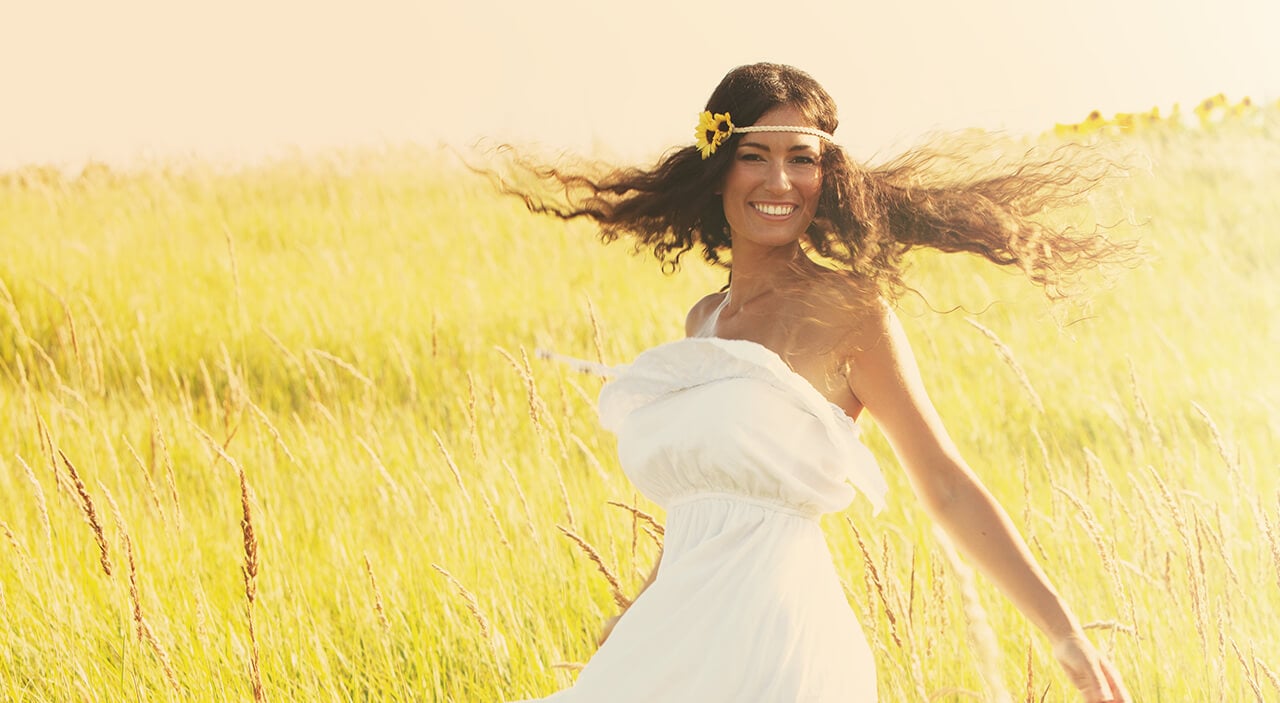Woman with long brown hair smiling wearing strapless dress and dancing in a grassy field.