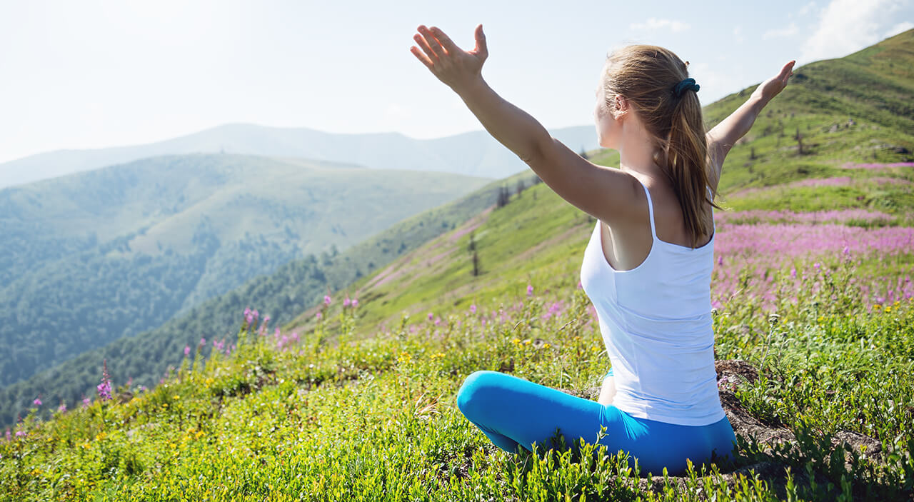 woman sitting on high mountain on the grass looking out with hands upward.