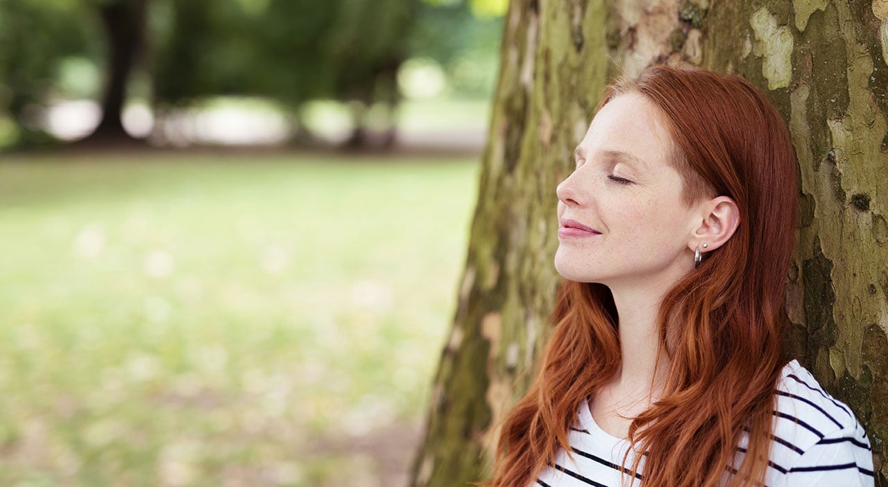Woman with eyes closed and head tilted up, smiling at the sky and leaning against a tree.