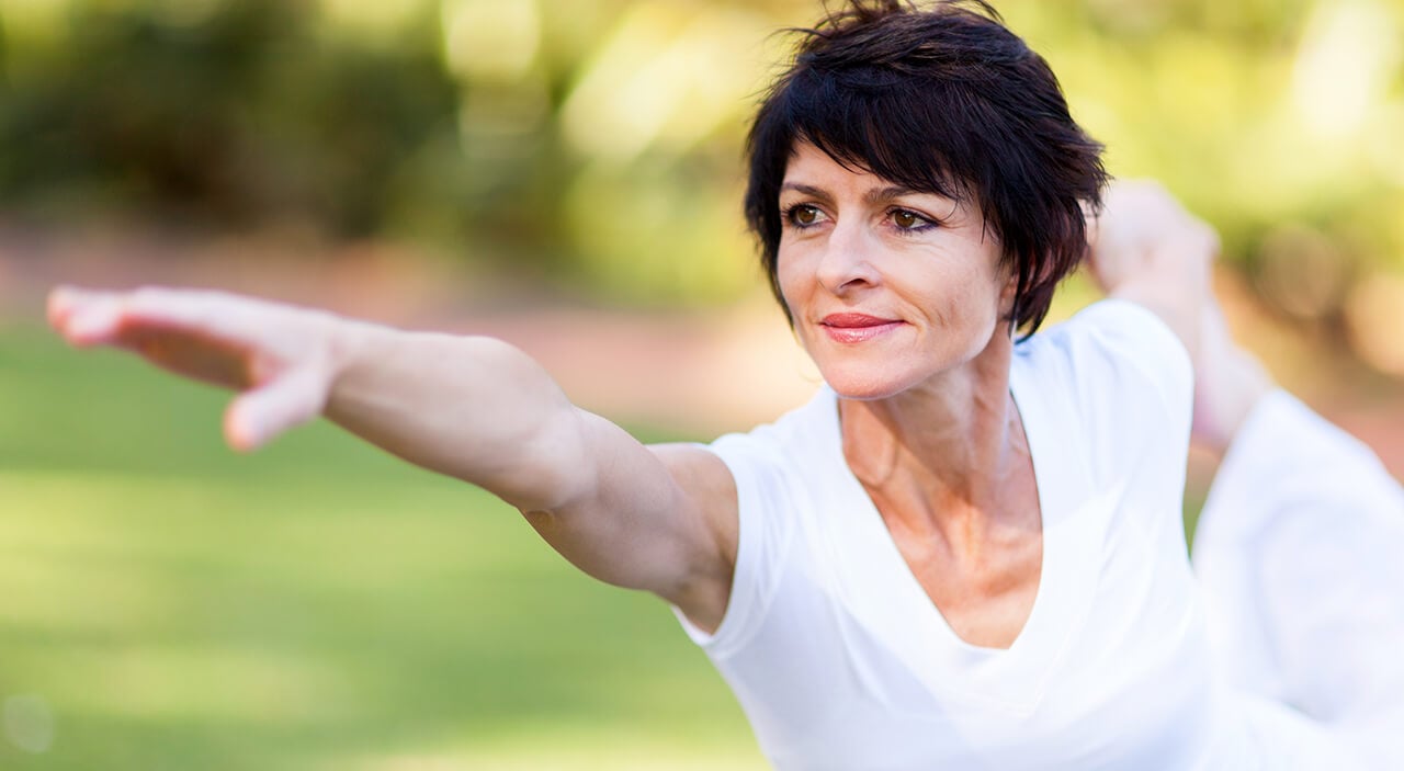 Older woman with short brown hair wearing white outfit holding a yoga pose with hand outstretched forward.