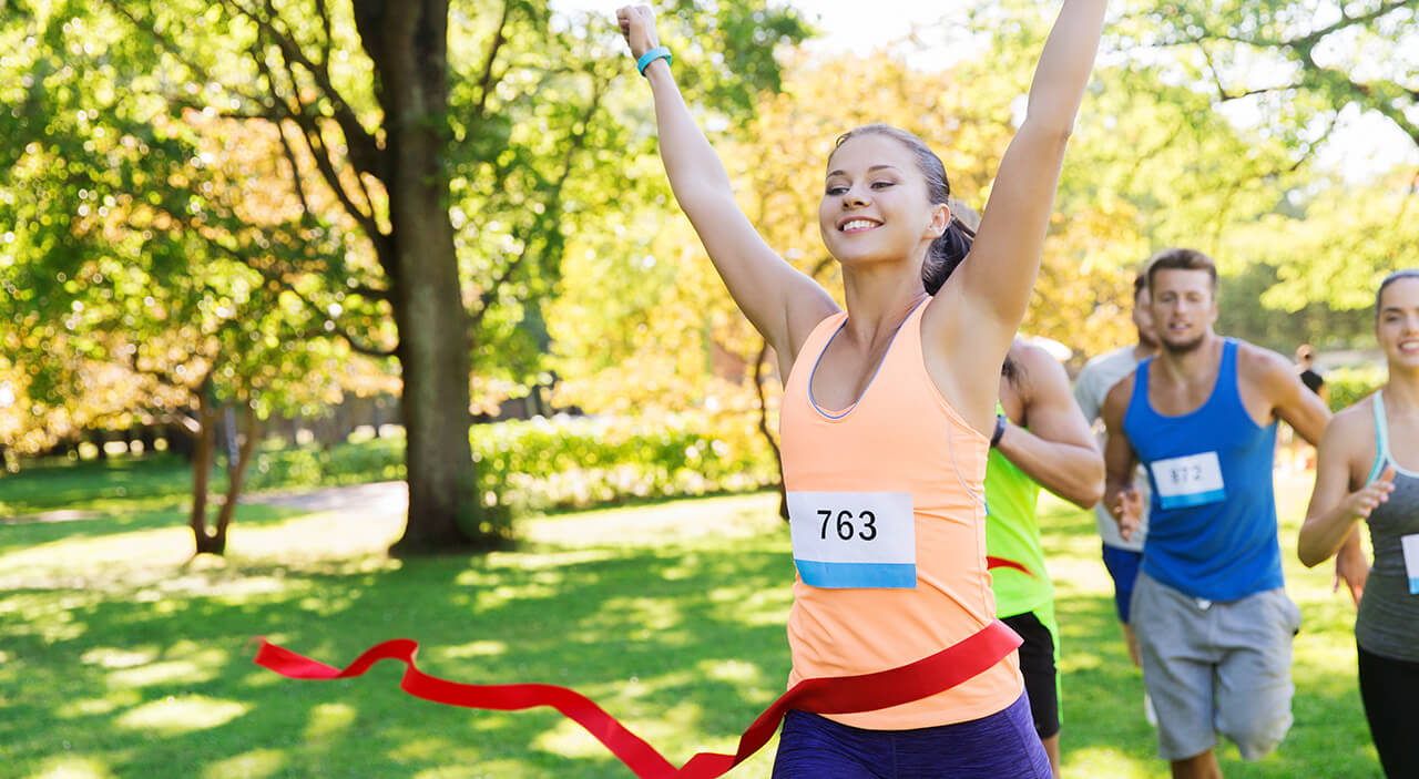 Young woman with brown hair in a ponytail smiling and running in a marathon with people around her.