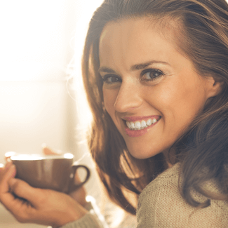 Woman wearing beige sweater and smiling as she drinks coffee.