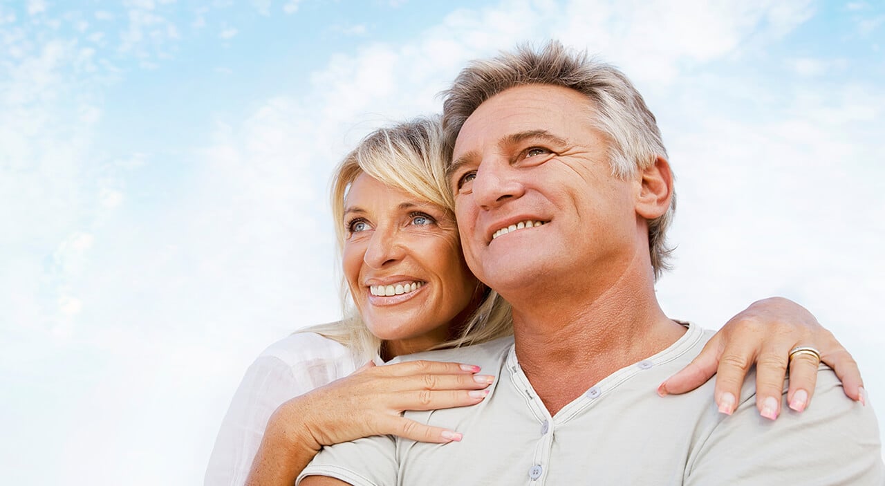 Older woman resting chin and hands on the shoulder of her husband as they both look out into the skin.