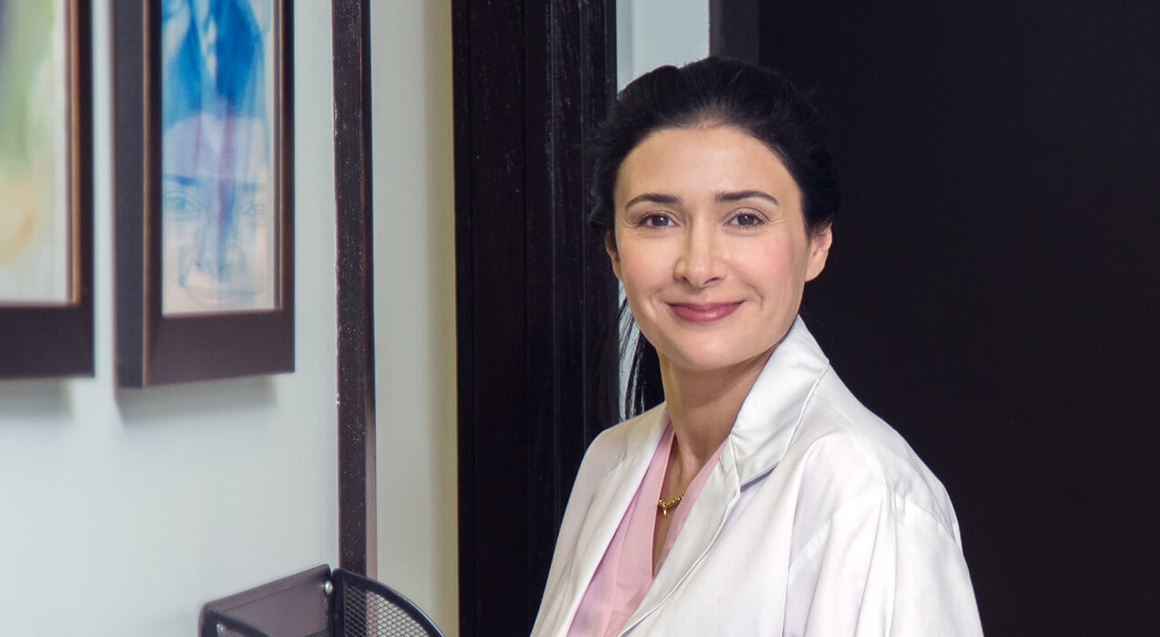 Toronto plastic surgeon Dr. Leila Kasrai in white lab coat and pink scrubs smiling.