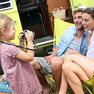 mother and father sitting on chairs as they watch their daughter take a picture while camping.