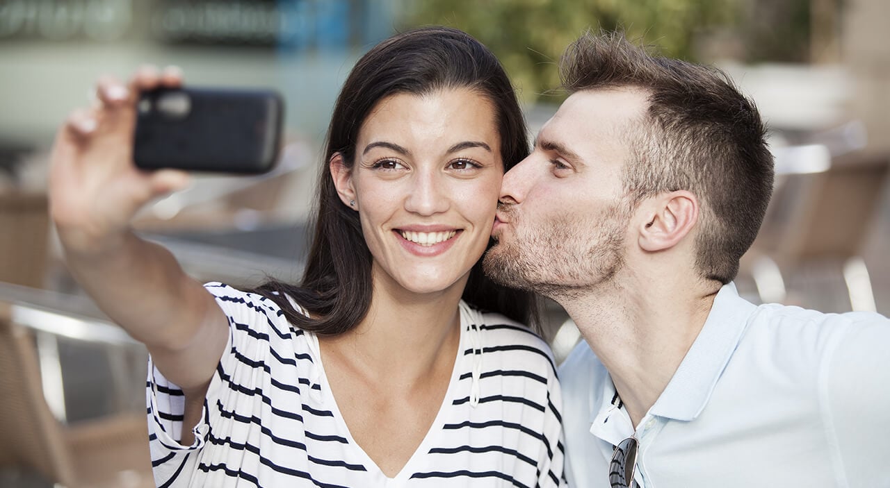 Woman with brown hair taking a selfie with her phone as her boyfriend kisses her on the cheek.