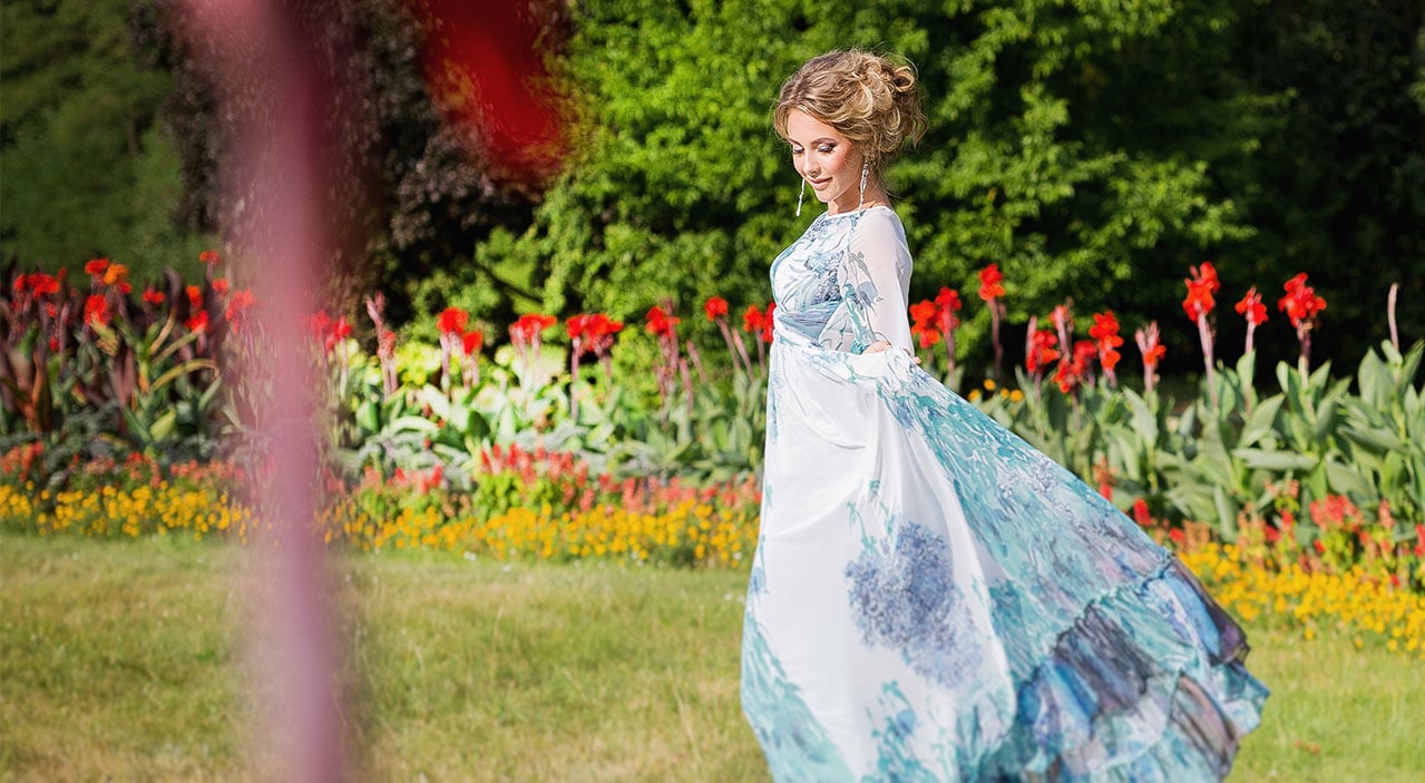 Young woman wearing long dress with hair looking down while standing in green garden with beautiful flowers.