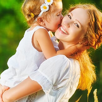 Mother holding daughter who is kissing her on the cheek who are both wearing white dresses in the park.