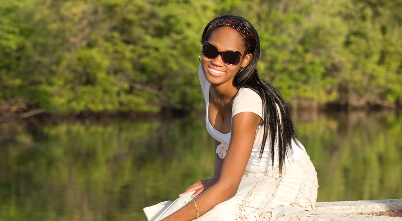 Woman sitting on dock at lake wearing white dress and sunglasses smiling.