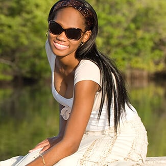 Woman wearing white dress and sunglasses smiling as she sits on a dock at a green lake.