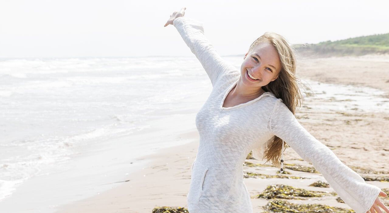 Young woman wearing white long sleeved shirt and smiling with hands extended out standing on the beach.