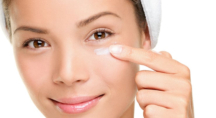 A smiling woman putting on cream on the skin under her eyes.